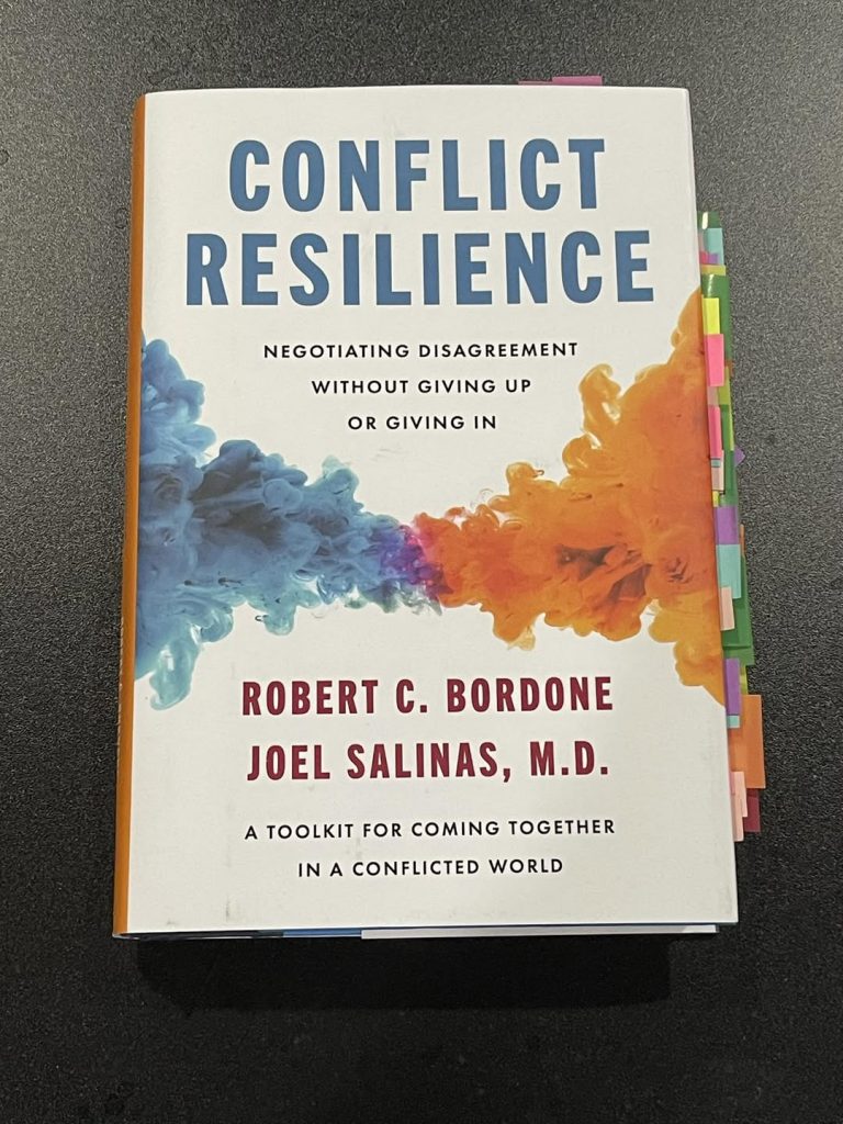 Book cover for Conflict Resilience: Negotiating Disagreement Without Giving Up or Giving In by Robert C. Bordone and Joel Salinas, M.D. 
The book cover has blue swirls meeting orange swirls in the middle, and there are many note tabs on the side.